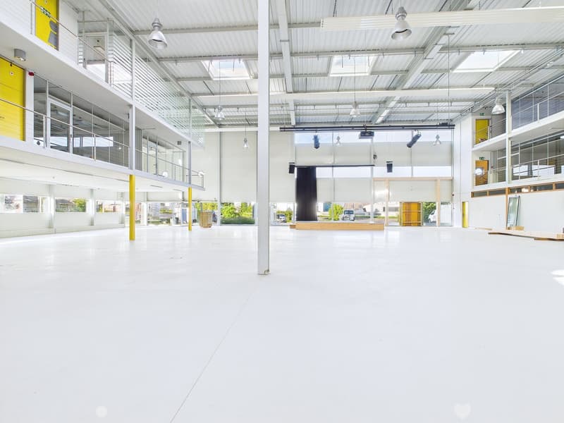 Large open commercial space with high ceilings, white walls, and yellow accents. The space has a stage-like area and appears to be a multipurpose event or exhibition hall.