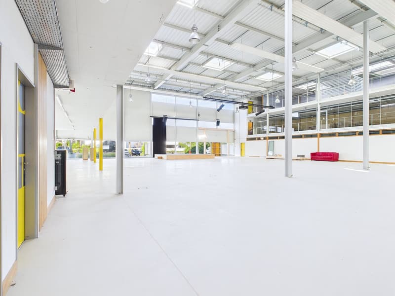 Large open commercial space with high ceilings, exposed ductwork, and plenty of natural light from the windows. The space has a clean, modern aesthetic with white walls and floors. There is a red sofa visible, suggesting the space may be used for events o