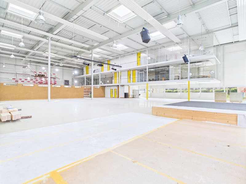 Large open warehouse space with high ceilings, white walls, and yellow accents. The space has a concrete floor with yellow markings, and there are various storage racks and equipment visible.
