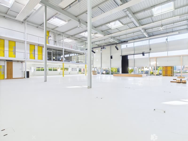 Large open commercial space with high ceilings, white walls, and yellow accents. The space has a stage area and appears to be set up for events or performances. There are also some storage areas and parking spaces visible.