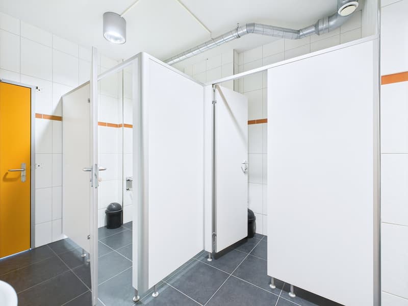 The image shows a modern bathroom with white walls, gray tile flooring, and glass shower stalls. The bathroom has a clean and minimalist design, with a ventilation system visible in the ceiling.