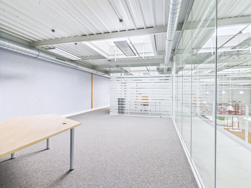 The image shows a modern, open-plan office space with a clean, minimalist design. The space features a large desk, glass walls, and a suspended ceiling with exposed ductwork. The floor is covered in a gray, speckled carpet. The overall aesthetic is bright