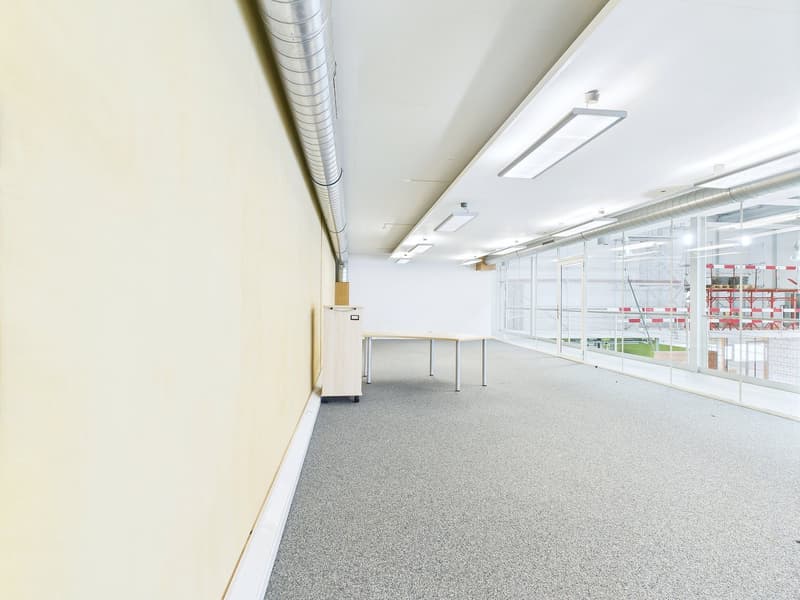 The image shows a large, open office space with white walls, a gray carpeted floor, and overhead lighting fixtures. There is a table visible in the center of the room, and the space appears to be empty and unoccupied.