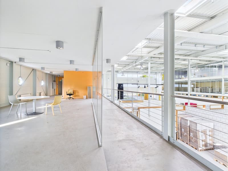 Large open-plan office space with concrete floors, white walls, and glass partitions. The space features a seating area with yellow chairs and a table, as well as a raised platform with additional seating. The ceiling is high and has exposed beams and lig