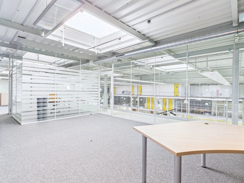 Large open office space with high ceilings, glass partitions, and a table in the foreground. The space appears to be well-lit and modern in design.
