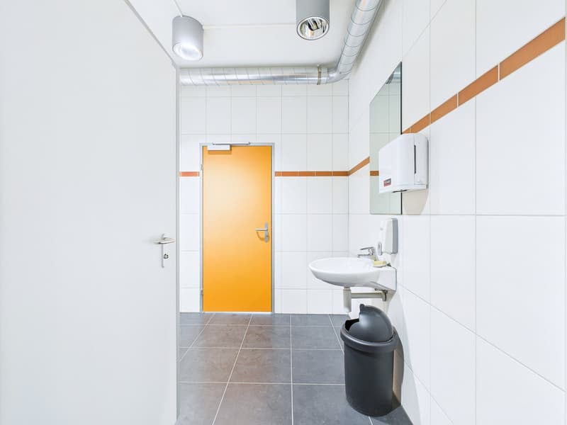 Tiled bathroom with a white sink, a black trash can, and an orange door. The room has white walls and gray floor tiles.