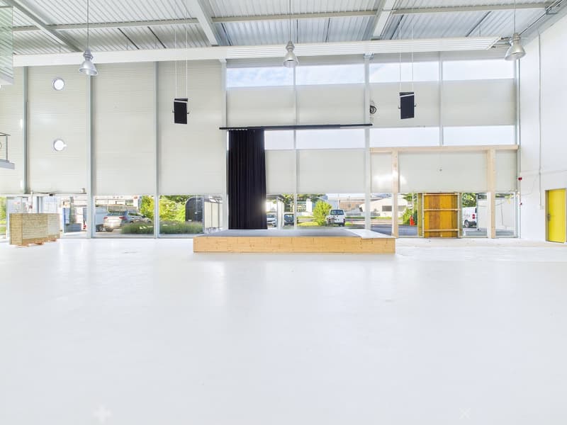 Large open commercial space with high ceilings, large windows, and a raised wooden platform in the center. The space appears to be used for events or presentations, with lighting fixtures and a curtain setup on the platform.