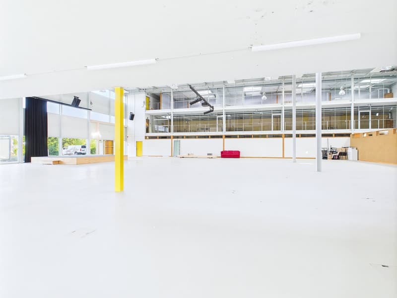 Large open commercial space with white walls, yellow support columns, and glass windows. The space appears to be empty and ready for use, with a red sofa visible in the background. There is a parking area visible through the glass windows.