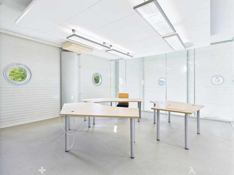 Bright and modern office space with large windows, white walls, and minimalist furniture including desks and chairs. The room has recessed lighting and a clean, professional atmosphere.