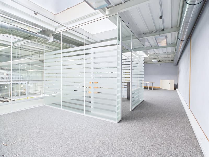 The image shows a modern, open-plan office space with glass partitions, a high ceiling, and exposed ductwork. The floor is covered in a gray carpet, and there are several chairs and a small table visible in the background.