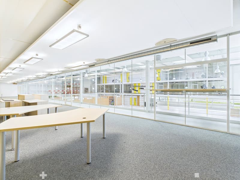 Large open office space with glass walls, desks, and shelving units. The space appears to be well-lit with overhead lighting and has a modern, minimalist design.