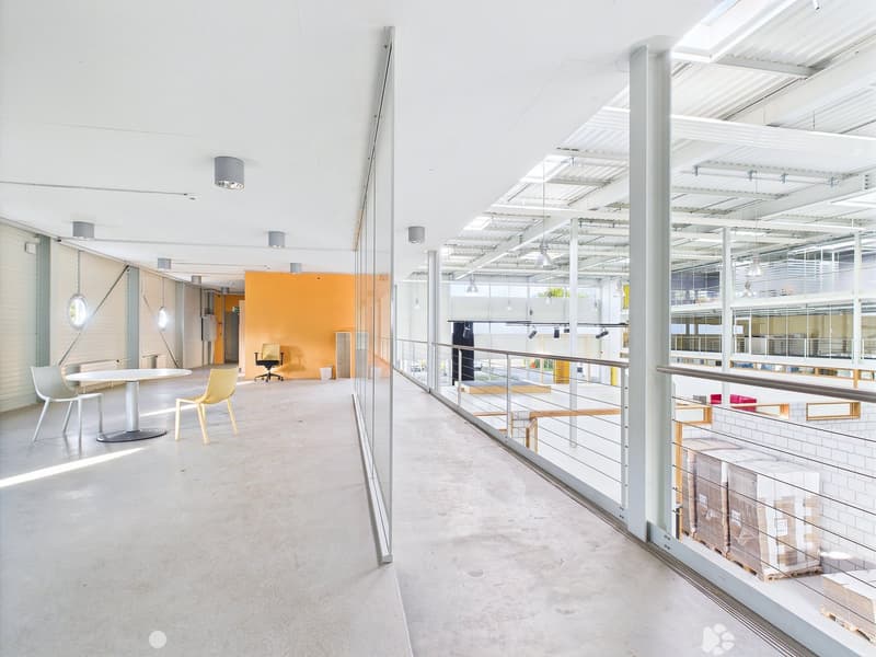 Large open-plan office space with concrete floors, white walls, and glass partitions. The space features a seating area with yellow chairs and a table, as well as a raised platform with additional seating. The ceiling is high and has exposed beams and lig