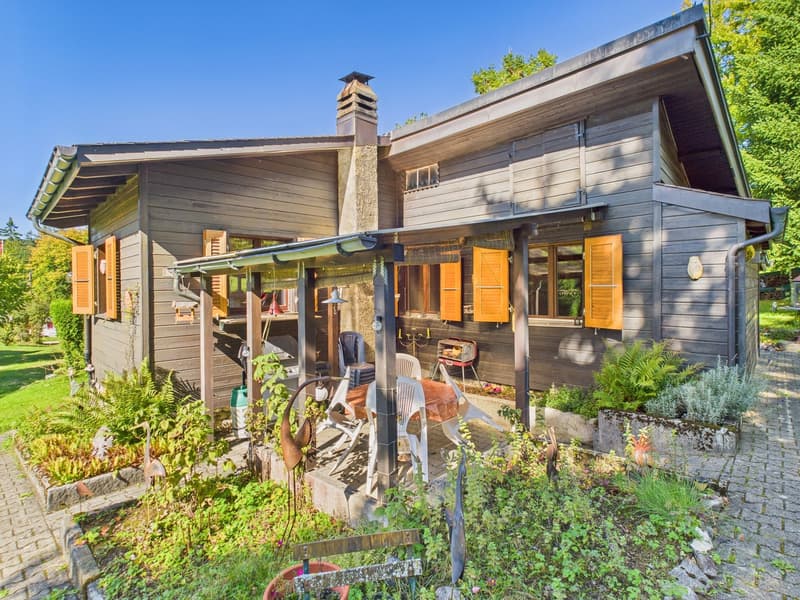 detached house, wooden exterior, yellow shutters, chimney, outdoor dining area with table and chairs, garden with various plants and shrubs, brick pathway, clear blue sky