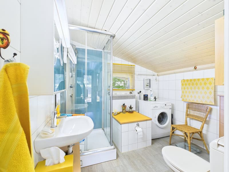 Small bathroom with a shower, toilet, sink, washing machine, small countertop with towel and a chair