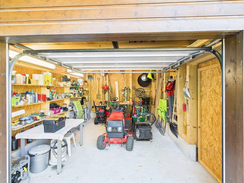 Interior of a garage, with storage shelves on the left, a workbench, and various tools and equipment including a red tractor and lawn mowers