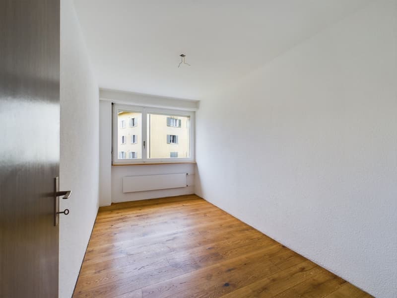 This image shows a simple, minimalist bedroom or living space. The room has white walls and a wooden floor. There is a window with a view of the exterior building. The room appears to be empty, with no furniture or decor visible.
