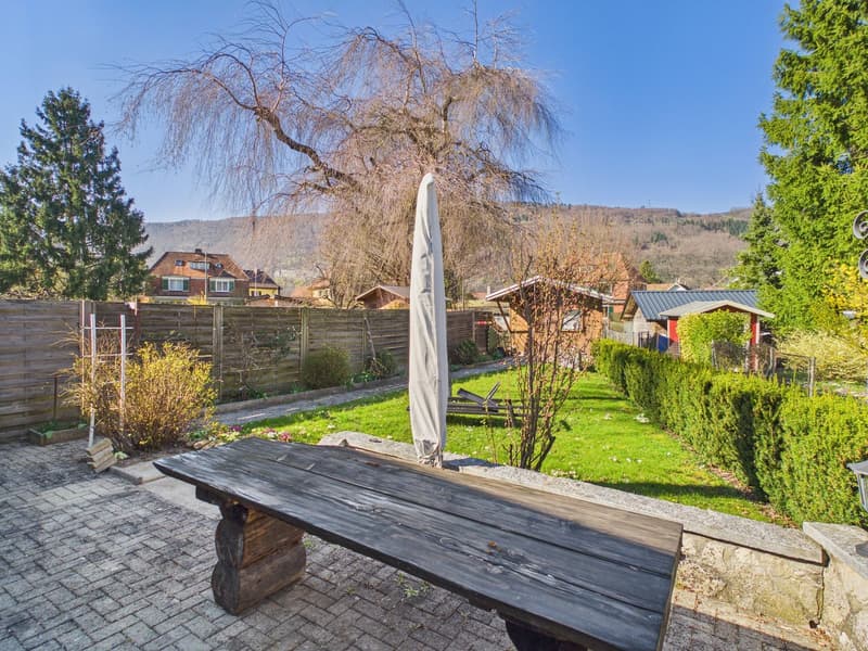 wooden table, closed umbrella, brick floor, wooden fence, green grass, trees, houses in the background