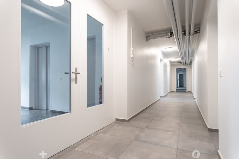 Empty hallway with white walls, tiled floor, glass doors, and a fire extinguisher
