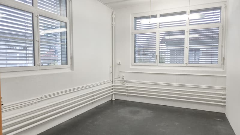 The room has two windows with blinds, a ceiling-mounted light fixture, and an electrical outlet on the wall. It is empty, with white walls, gray floors, and pipes on the floor.