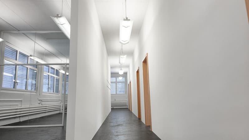 a modern hallway with white walls and grey floor. Glass partitions on the left, doors on the right.