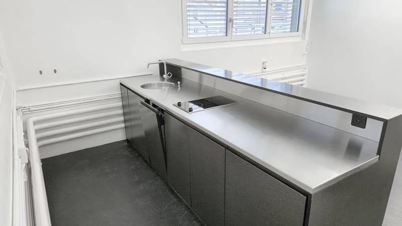 The room features a stainless steel countertop with an oven below, a sink, a faucet, and three cabinets. There is also a window with blinds and a radiator.