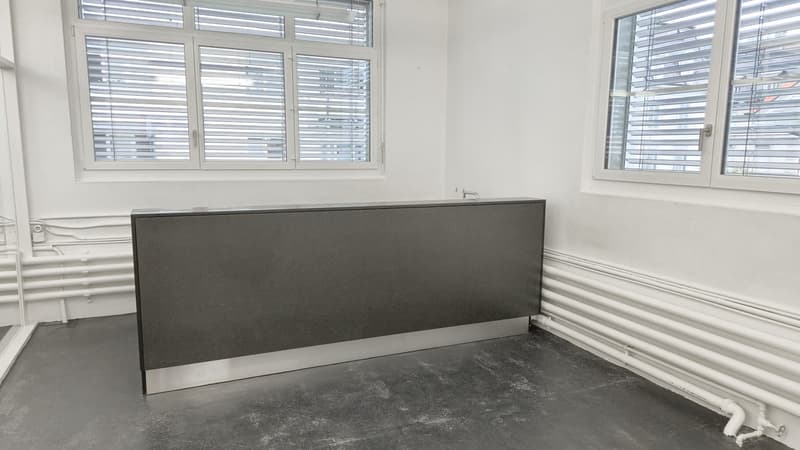 modern office with white walls, gray floor, desk with metal base, radiators, two large windows with blinds, pipes on wall