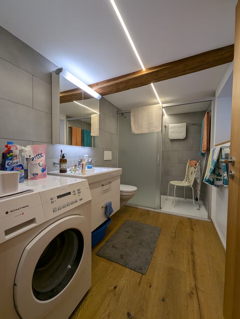Bathroom with a washing machine, vanity with sink, mirror, and shelving. Wooden beams on the ceiling and hardwood flooring.