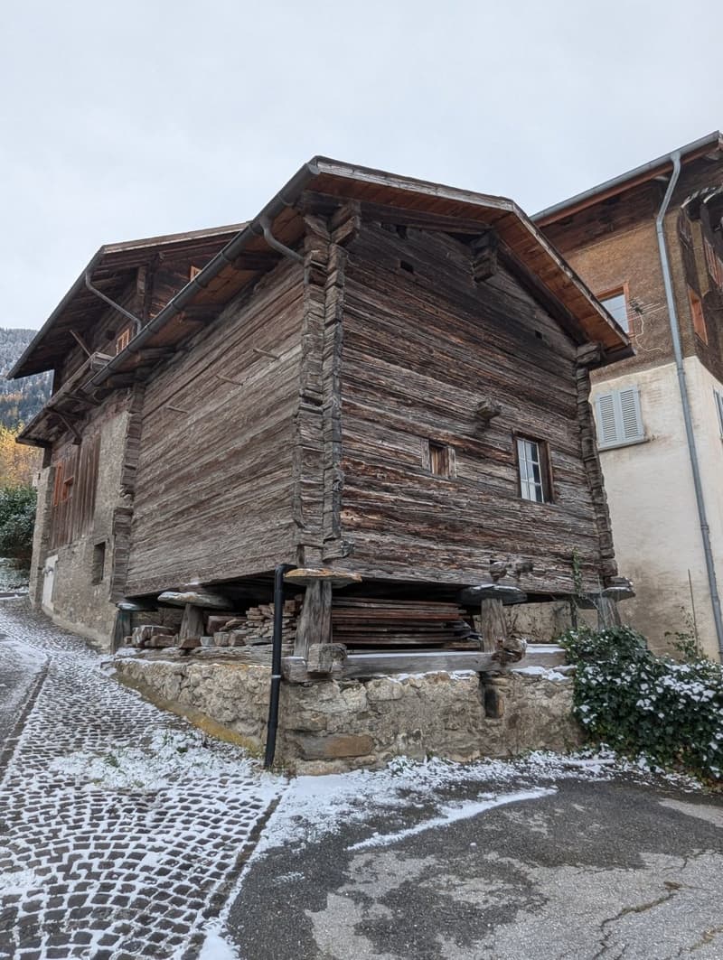 Old, rustic wooden house with a traditional alpine architecture style. The house has a steep, pitched roof and is constructed using large, weathered wooden logs. The exterior walls are made of wooden planks and the building appears to be situated on a cob