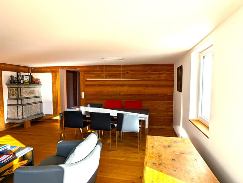The image shows a cozy and rustic open-plan living and dining area. The walls are made of wooden panels, creating a warm and inviting atmosphere. There is a fireplace visible, which adds to the coziness of the space. The dining table is positioned in the