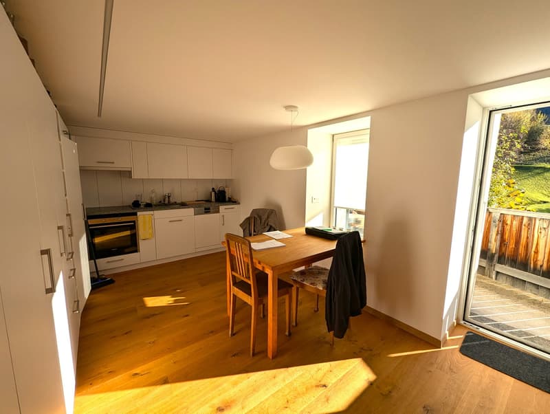 The image shows a combined kitchen and dining area with a wooden dining table and chairs, a modern kitchen with white cabinets and appliances, and a large window overlooking a garden or outdoor area.