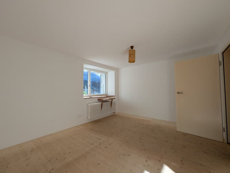 This image shows an empty room with white walls, a wooden floor, and a radiator under the window. The room appears to be a bedroom or living space, with a simple light fixture hanging from the ceiling.