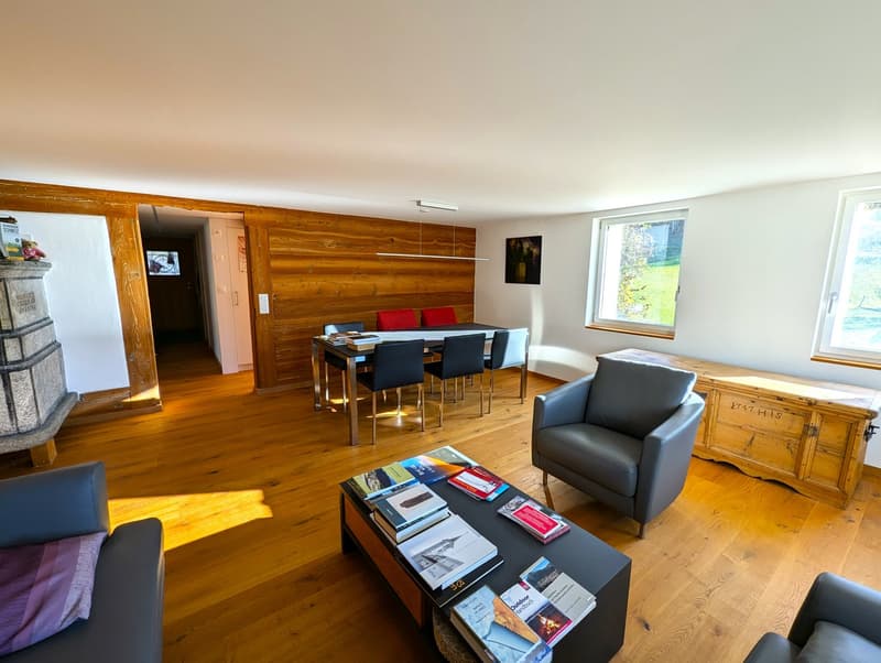 Spacious living room with wooden walls, hardwood floors, large windows, dining table, sofa, armchair, and a fireplace