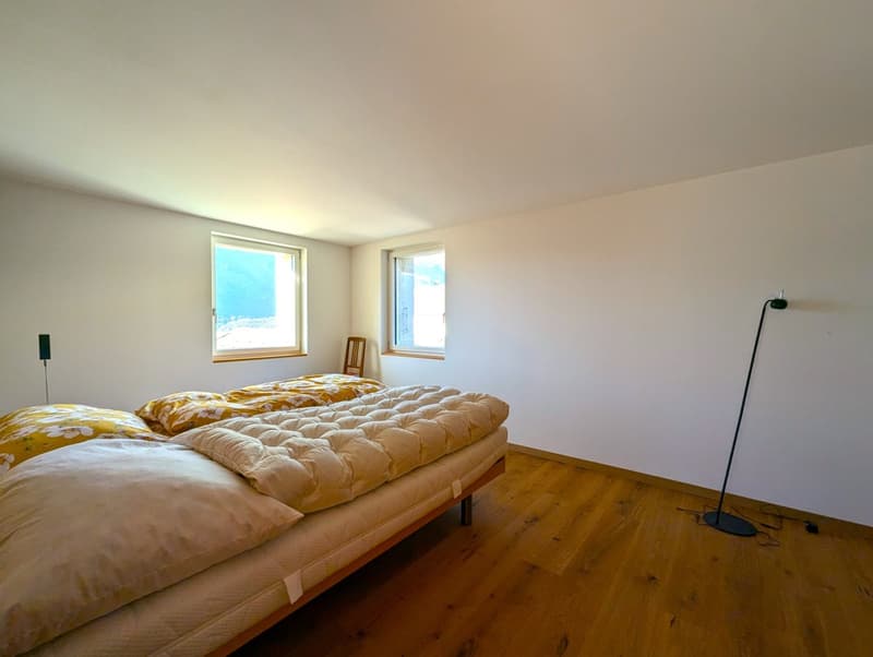 Spacious bedroom with large windows, hardwood flooring, and a comfortable bed with floral bedding. The room has a cozy and bright atmosphere.