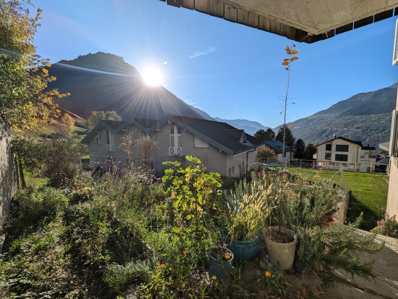 Two-story house with a slanted roof, surrounded by a lush garden with various plants and flowers. The house is situated in a mountainous area with a clear blue sky and sunlight shining through.