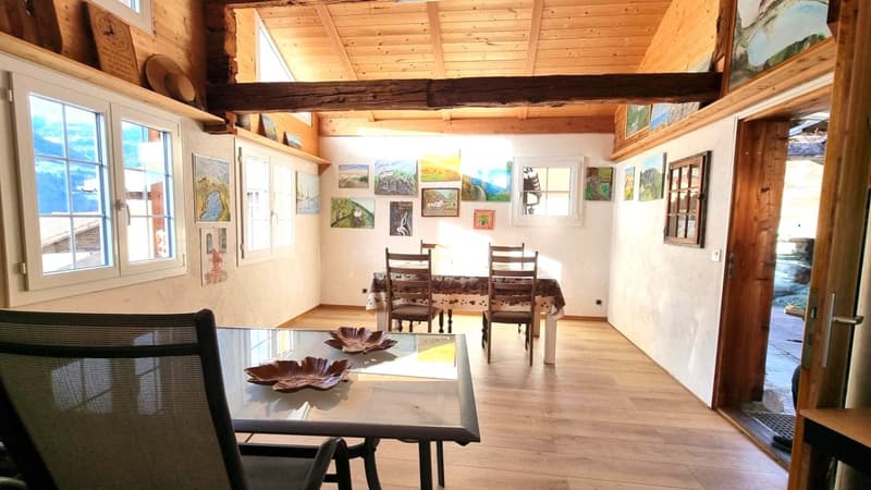 Spacious open-plan living and dining area with wooden beamed ceiling, large windows, wooden floors, and artwork displayed on the walls. The room features a dining table with chairs, and a fireplace is visible in the corner.