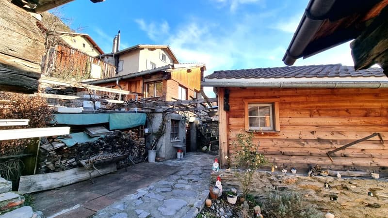 Two-story wooden house with a stone foundation, a garage, and a parking area. The house has a tiled roof and appears to be in a rural or mountainous setting with trees and other buildings visible in the background.