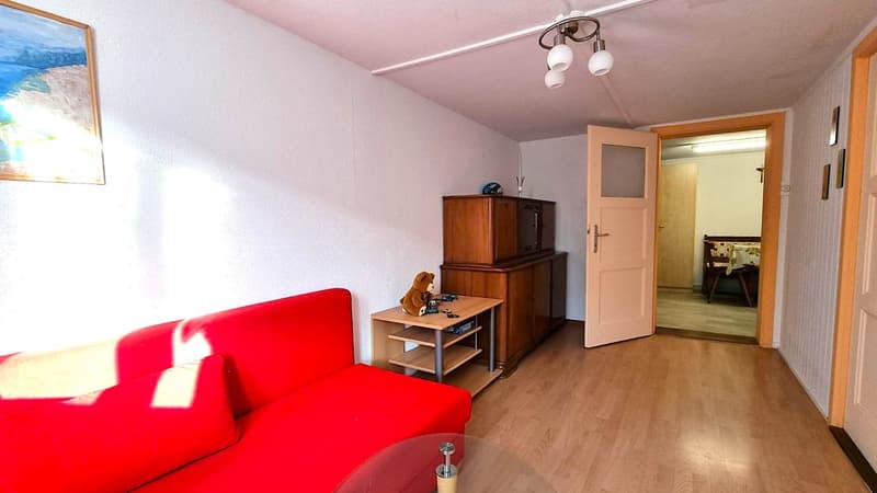 Spacious living room with a red sofa, wooden furniture, and a door leading to another room. The room has a hardwood floor and a ceiling light fixture.