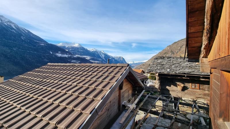 The image shows a traditional wooden mountain chalet with a tiled roof, situated in a scenic alpine landscape. The chalet is surrounded by snow-capped mountains and a clear blue sky, providing a stunning view from the property.