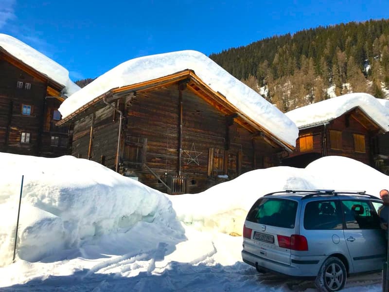 Wooden chalet in snowy environment, covered in snow, three windows, brown wooden doors, SUV parked in front