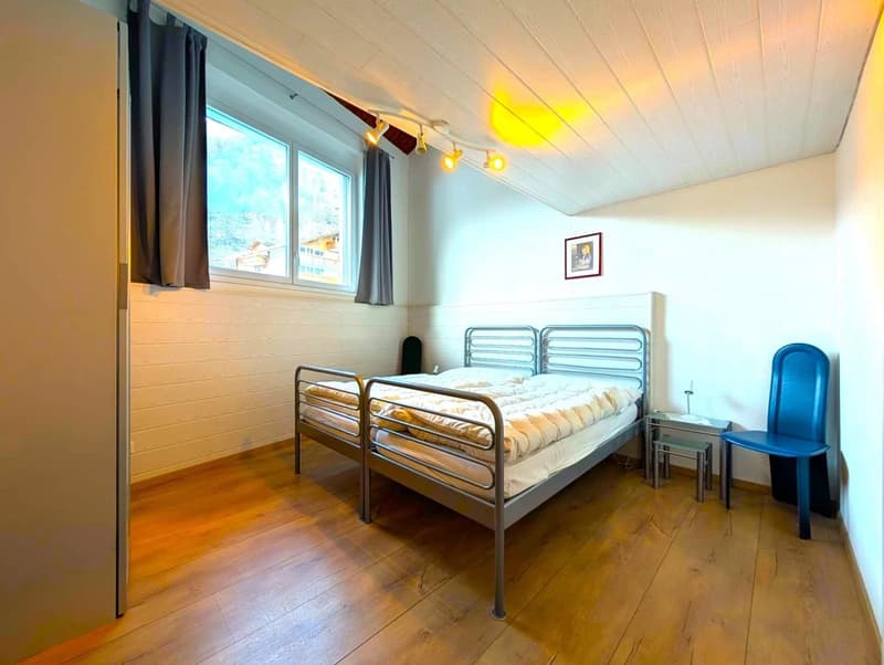 Two single beds, bedside table with lamp, blue chair, ceiling spotlights, wooden floor, white walls, window with curtains, picture frame