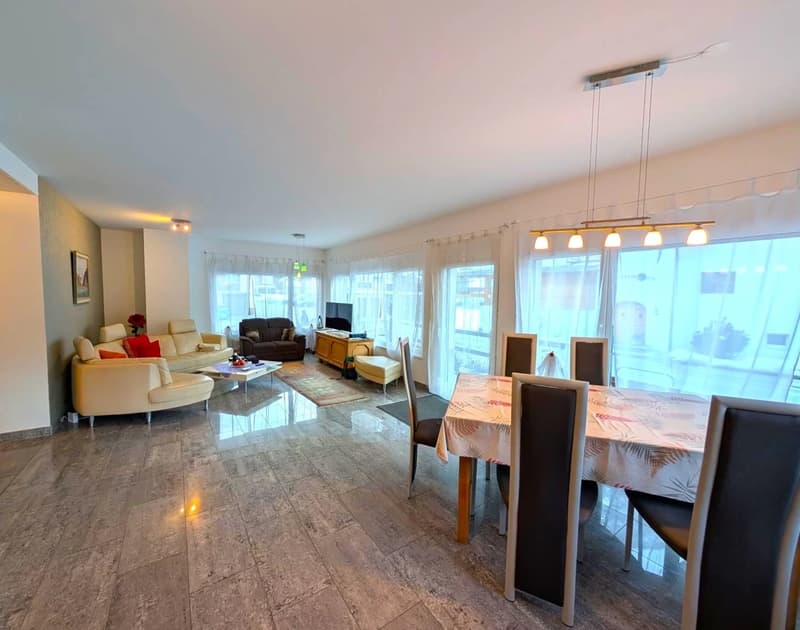 Spacious open layout with a living room area featuring a sofa set, a coffee table, and a TV. Dining area with a dining table and chairs. Wooden floor, white walls, and large windows. No visible visible features in the room.