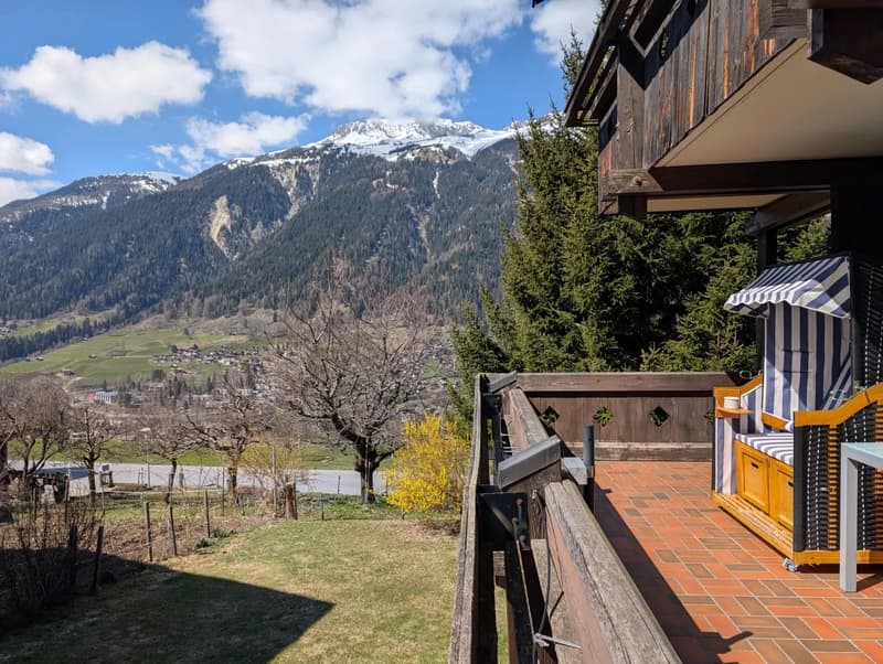 2-story wooden chalet-style building with a balcony and terrace overlooking a scenic mountain landscape with snow-capped peaks, forested hills, and a village in the valley below