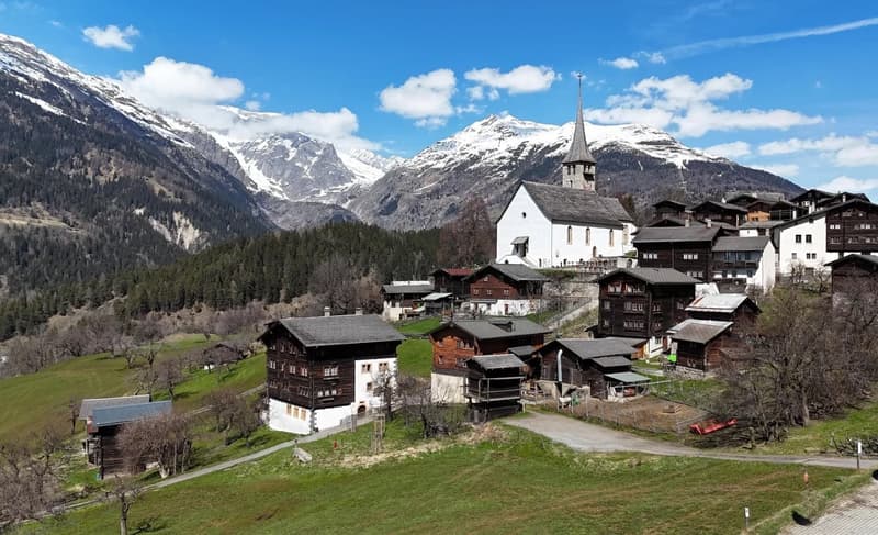Traditional alpine village with wooden chalets, set against the backdrop of snowy mountains, one with a steeple.