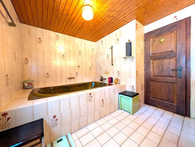 wooden ceiling, white tiled walls with floral patterns, bathtub, tiled floor, wooden door, faucet, wooden bench