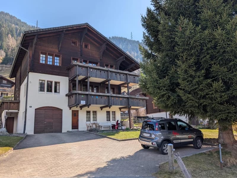 3-story chalet, wooden exterior, large windows, brown garage door, wooden balcony, grassy yard, car parked in front, mountains in the background