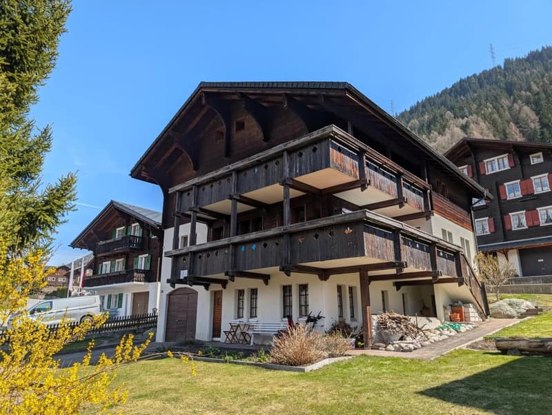 Traditional wooden architecture, multi-story, white and brown walls, large wooden balcony, white chairs, flags, driveway, mountain backdrop