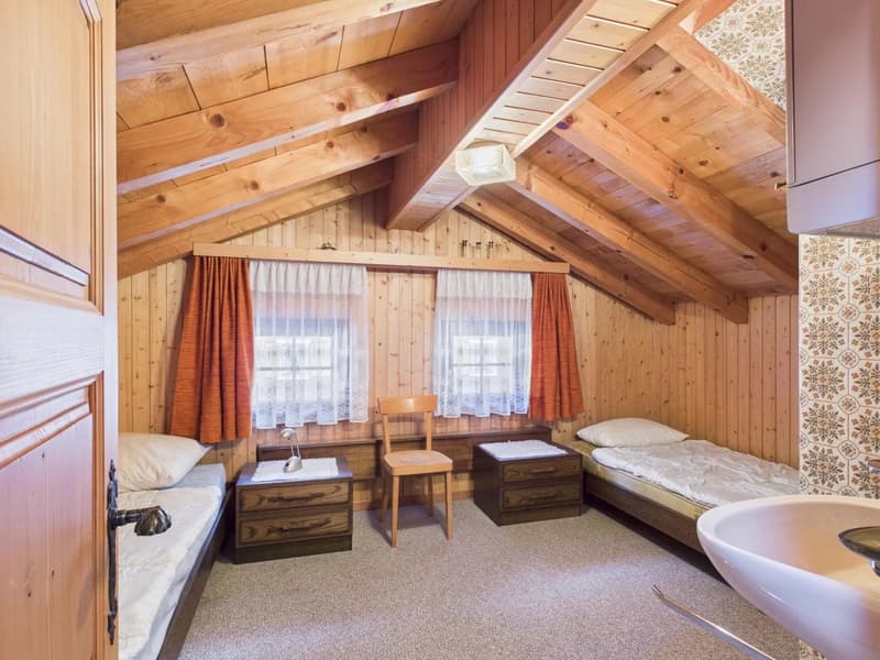 wooden walls, sloping ceiling, two single beds, wooden nightstands with lamps, small chair, windows with white curtains and red drapes, sink, no decorations