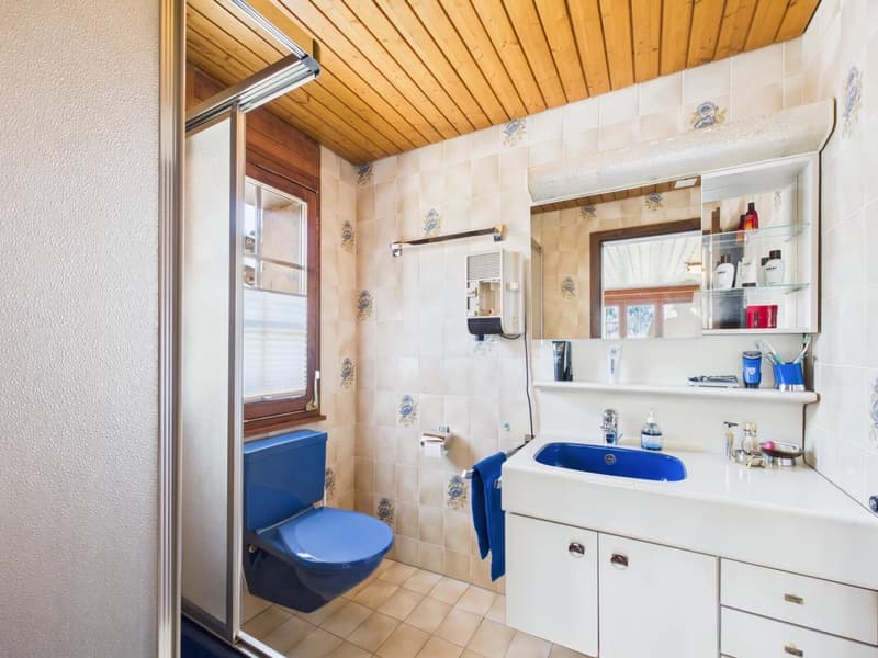 Bathroom with white tile walls, wooden ceiling, blue ceramic toilet, blue sink, white countertop, vanity with drawers, towel hanging on the wall, and various toiletries on the shelf.