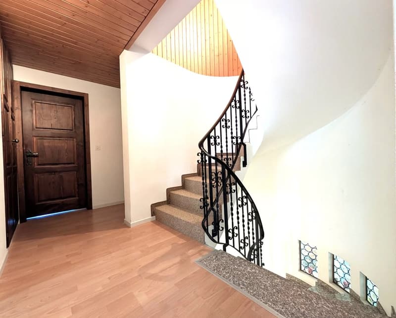 Spiral staircase with a black railing, wooden floors, white walls, a wooden door with a blue light strip at the bottom, decorative stained glass windows on the wall below the stairs