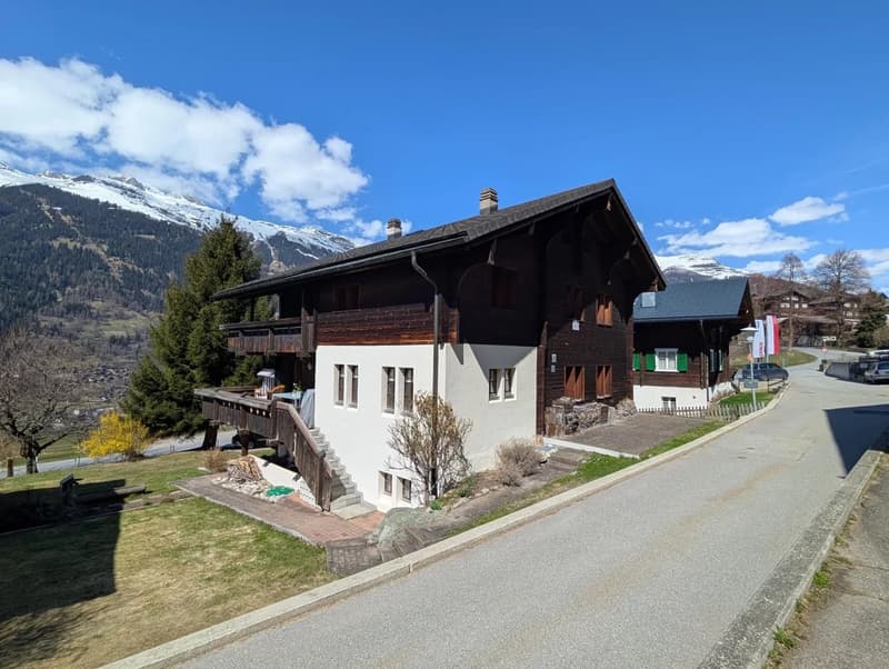 2 story chalet-style building with wooden and white exterior, balconies, surrounded by mountains and trees in a scenic alpine setting